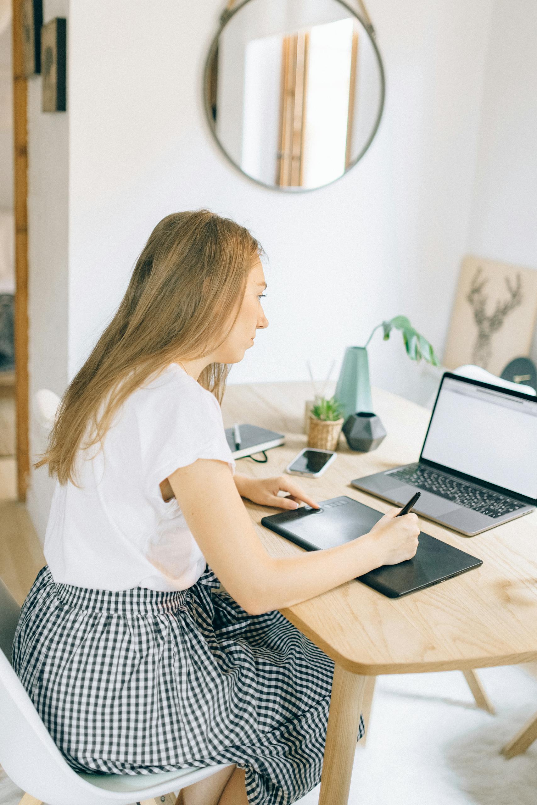 Young woman multitasking with laptop and graphic tablet in home office setting.